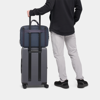 NOMATIC Laptop Bag in navy, worn cross-body next to rolling suitcase